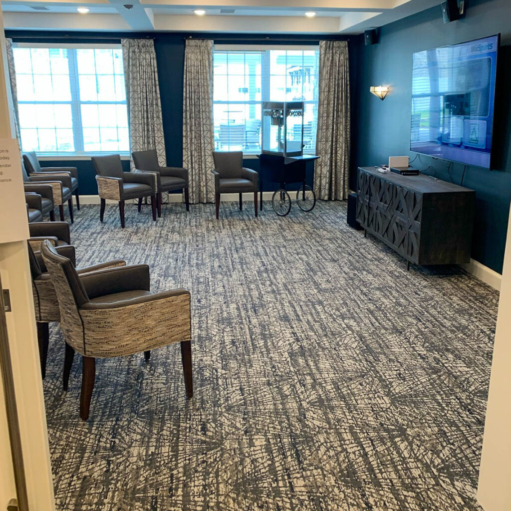 Theater room at the Boulevard of Saint Peters Senior Living, featuring comfortable chairs, a large TV, a popcorn machine, and large windows with curtains.