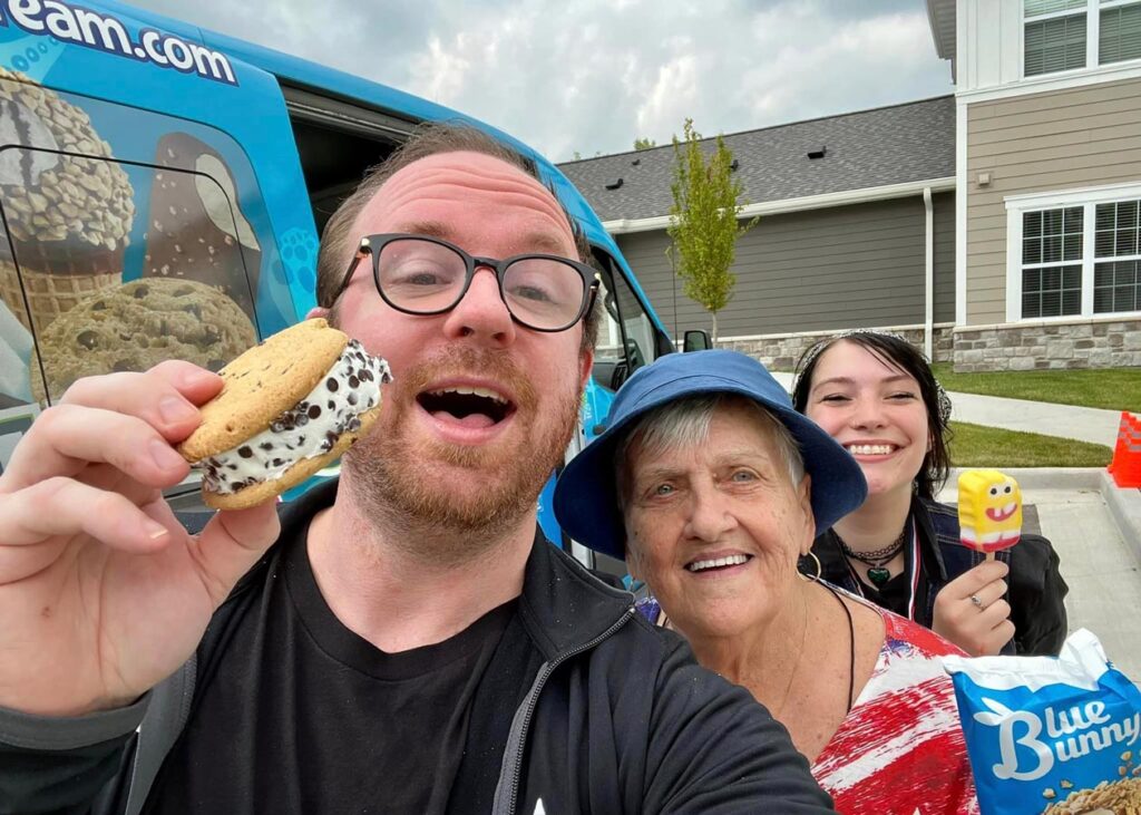 Three smiling individuals, including a senior woman in a blue hat, enjoy ice cream treats in front of a colorful ice cream truck. One person holds an ice cream sandwich, another holds a character popsicle, and a bag of Blue Bunny treats is visible, capturing a fun outdoor moment.