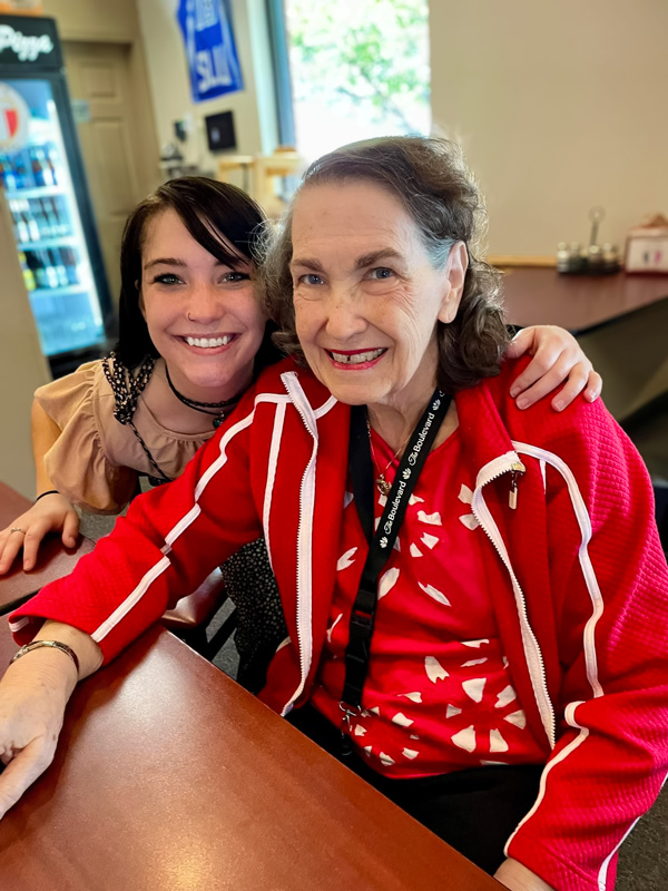 Senior resident and young woman smiling together at a table during an outing, with the resident wearing a red jacket and both sharing a moment of warmth and connection.